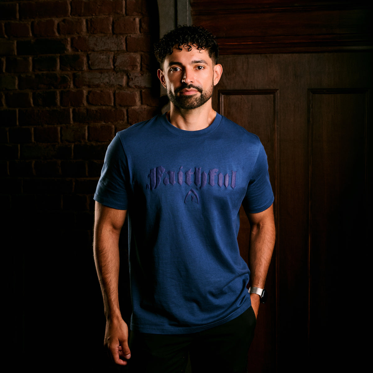 A man wearing a blue short-sleeve t-shirt with a dark gothic-style 'Faithful' graphic on the chest, standing in front of a dark brick wall and wooden door.