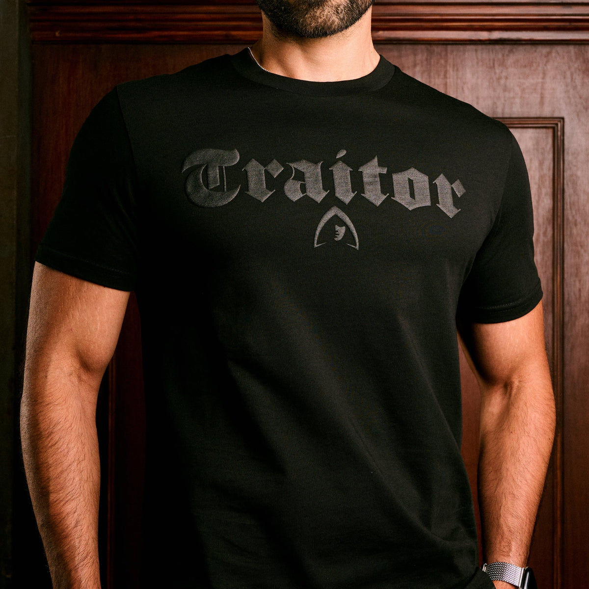 A man wearing a black short-sleeve t-shirt with a dark gray gothic-style 'Traitor' text graphic and a small shield emblem on the chest, set against a dark wood-paneled background.