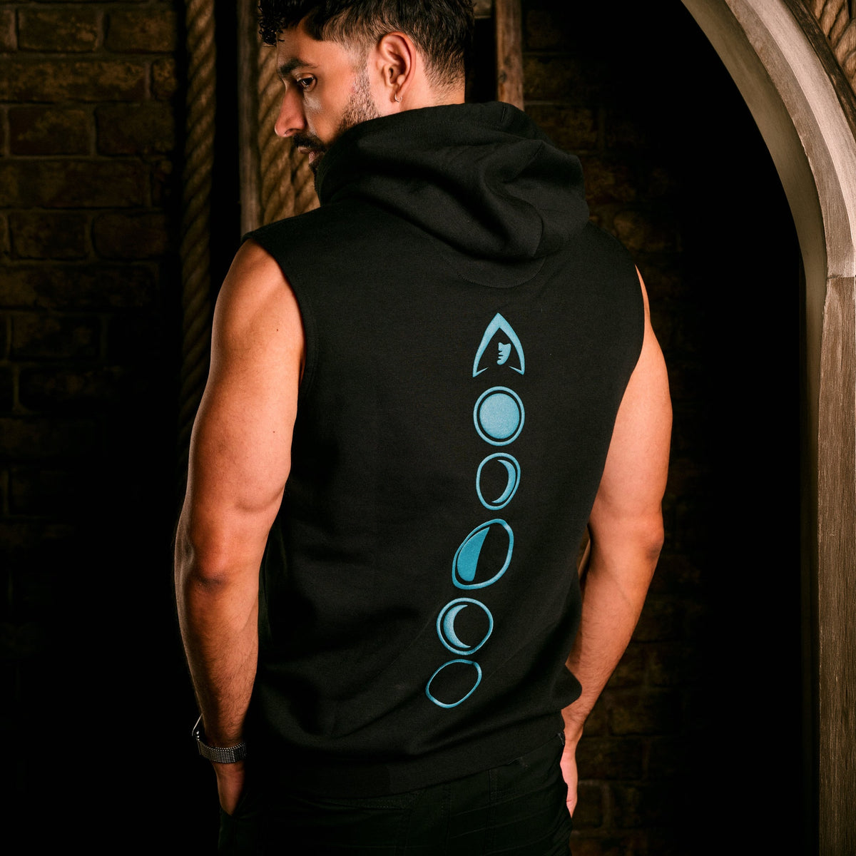 A man wearing a sleeveless black hoodie with a vertical moon phase graphic in light blue on the back, photographed in a dimly lit stone archway setting.