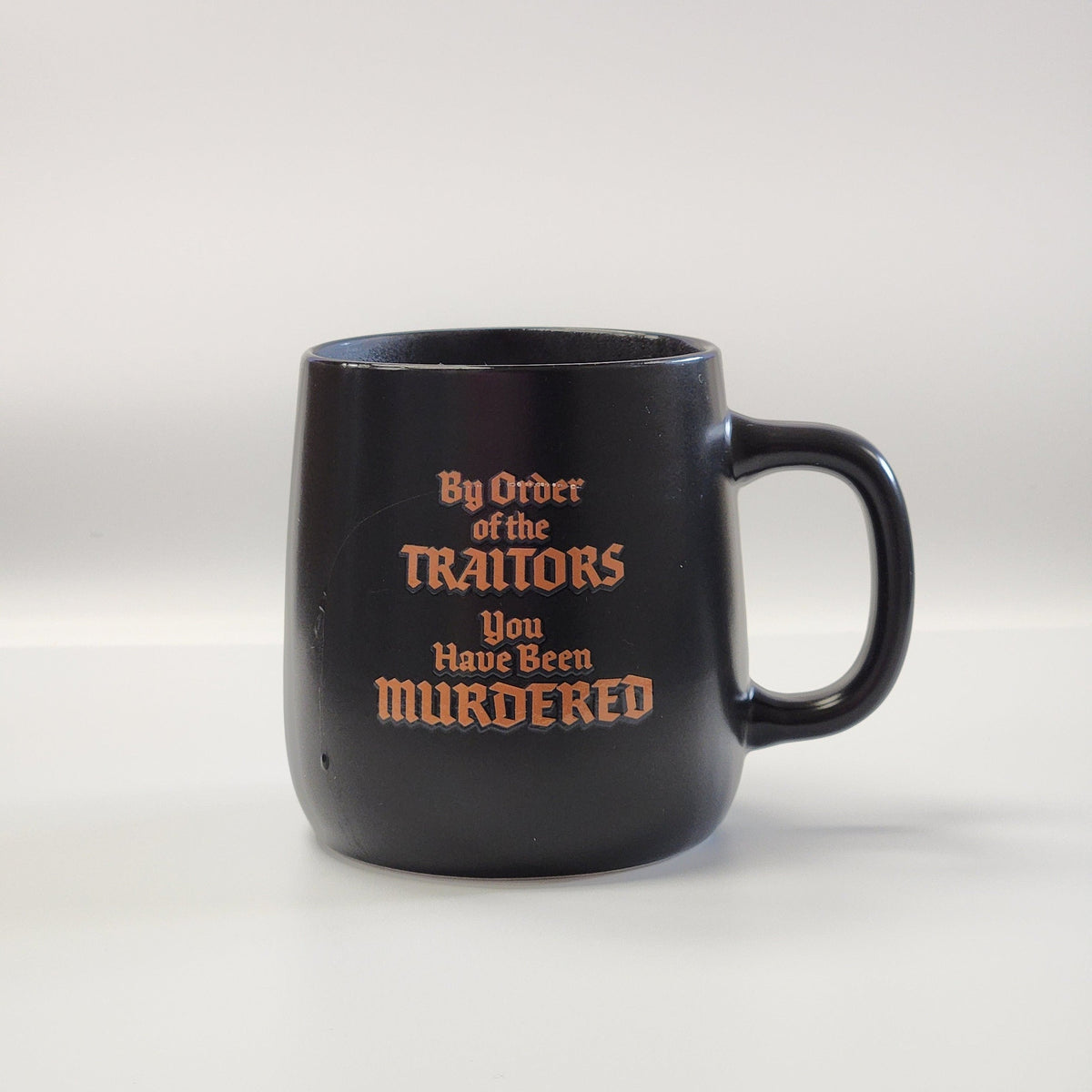 A black ceramic mug with a handle, featuring the text 'By Order of the TRAITORS You Have Been MURDERED' printed in a reddish-brown gothic font, set against a plain light gray background.