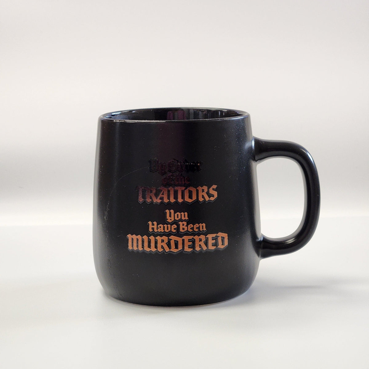 A black ceramic mug with a printed gothic-style text design reading 'Betrayals You Have Been MURDERED' in orange and dark lettering, set against a plain light gray background.