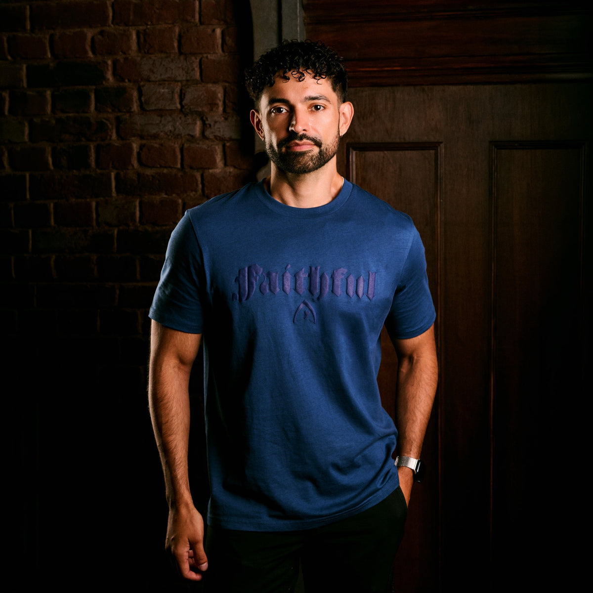 A man wearing a blue short-sleeve t-shirt with a dark gothic-style 'Faithful' graphic on the chest, standing in front of a dark brick wall and wooden door.