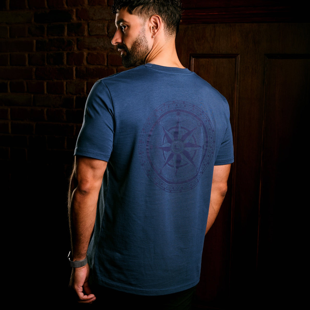 A man wearing a heathered blue t-shirt, viewed from behind to show a large circular compass rose graphic printed on the back, set against a dark brick wall and wooden door background.