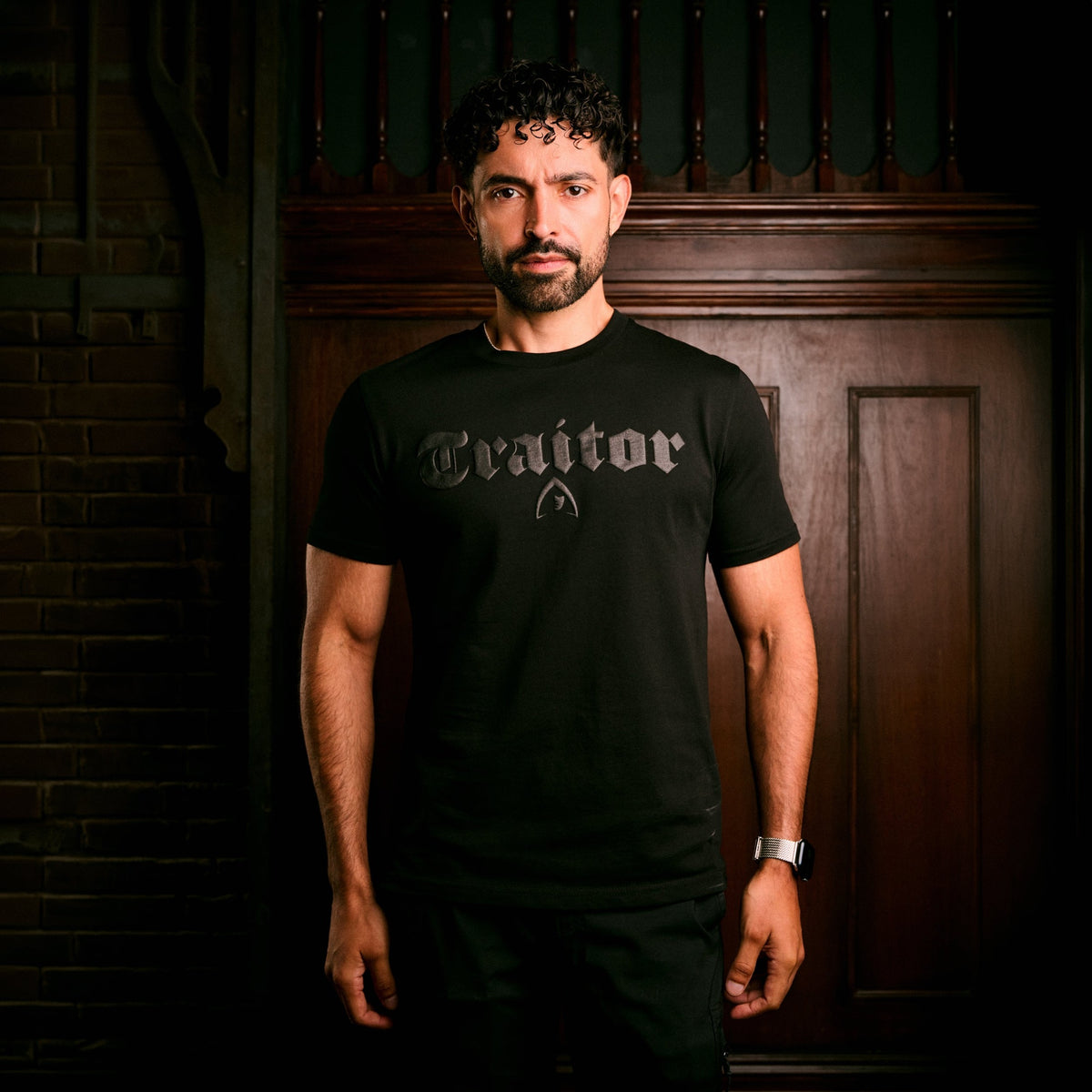 A man wearing a black crew-neck t-shirt with a gothic-style 'Traidor' graphic on the chest, standing in front of a dark wood-paneled wall.