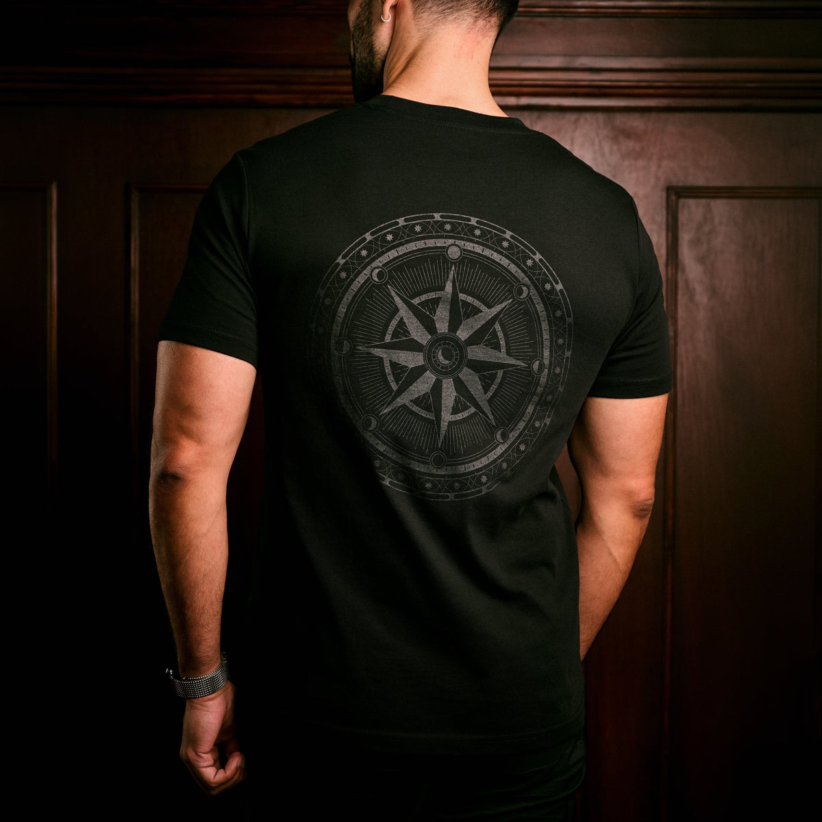 A man viewed from behind wearing a black t-shirt with a large gray compass graphic on the back, standing in front of a dark wood-paneled wall.
