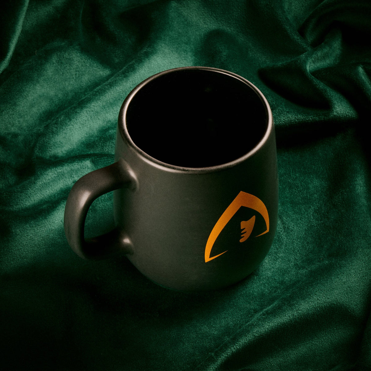 A matte black ceramic mug with a gold shield logo featuring a harp, resting on a draped dark green velvet fabric background.