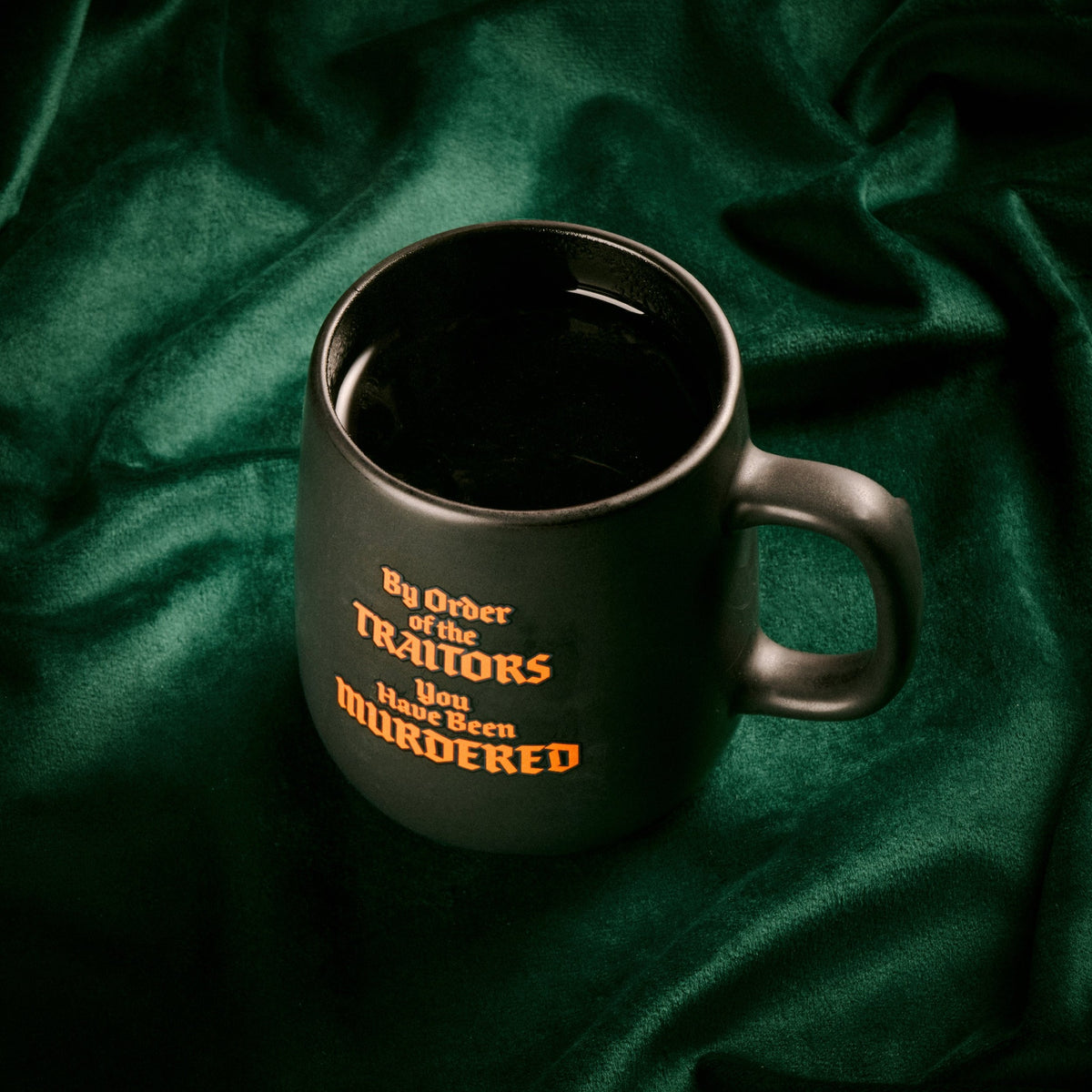 A dark gray ceramic mug with a handle, filled with black coffee, featuring the text 'By Order of the TITANS You Have Been MURDERED' in orange and yellow lettering. The mug is set against a draped dark green satin fabric background.