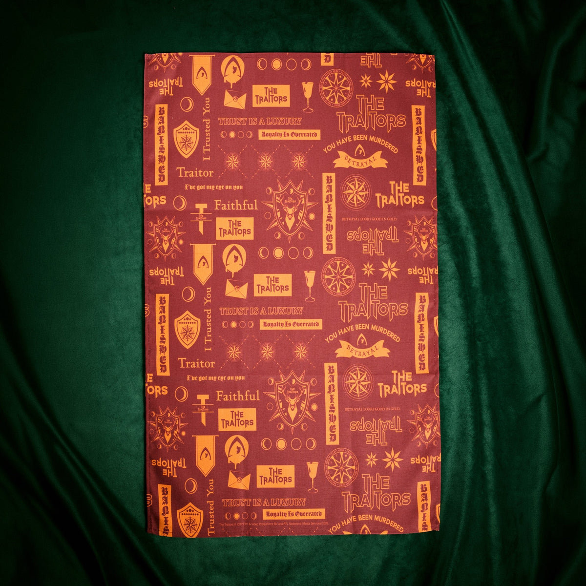 A rectangular red fabric item, likely a tea towel or scarf, featuring a repeating gold pattern of occult symbols, shields, and the words 'The Traits' and 'Faithful', displayed flat against a dark green satin background.