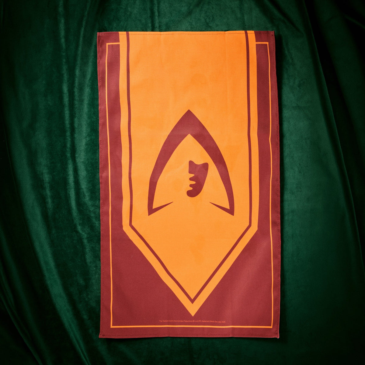 A rectangular flag or banner laid flat on a dark green velvet fabric background. The flag has a warm orange center with a large dark red geometric emblem and border details.